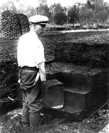 A man cutting peat. Note that the man is wearing puttees, not Wellington boots. Puttees were worn by agricultural workers together with hobnail boots, but were eventually made obsolete by cheap rubber boots.